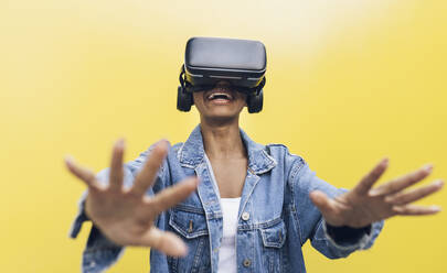 Smiling young woman wearing virtual reality headset gesturing against yellow background - JCCMF05150