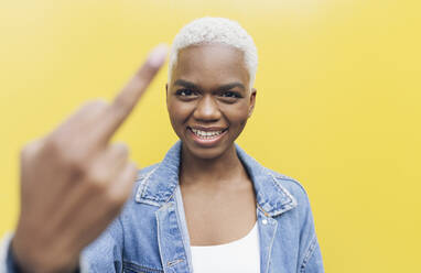 Smiling woman showing middle finger against yellow background - JCCMF05154
