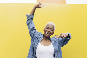 Happy woman making peace sign gesture against yellow background - JCCMF05158