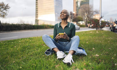 Smiling woman with smart phone sitting on grass in city - JCCMF05182