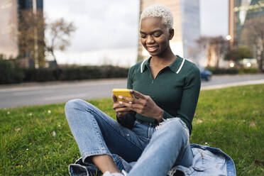 Junge Frau, die ein Smartphone benutzt, sitzt im Gras am Straßenrand - JCCMF05183
