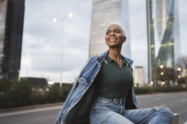 Smiling woman sitting at roadside in city - JCCMF05185