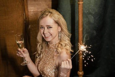 Cheerful young blond woman holding champagne flute and sparkler at New Year's party - SSGF00425