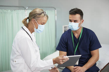 Ärztin mit Gesichtsschutzmaske bespricht sich mit Krankenschwester über Tablet-PC in medizinischem Raum - PMF02128