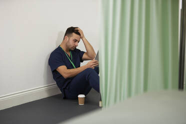 Besorgter Krankenpfleger mit dem Kopf in der Hand sitzt an der Wand in einem medizinischen Raum - PMF02136