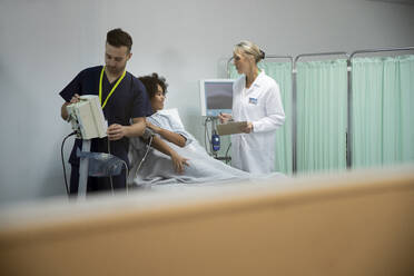 Krankenschwester, die ein Blutdruckmessgerät bedient, mit einem Patienten und einem Arzt, die sich im Hintergrund unterhalten - PMF02194