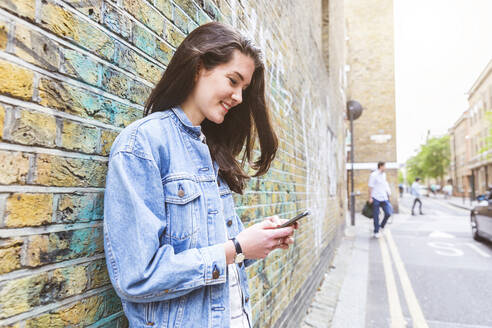 Lächelnde junge Frau, die ein Smartphone benutzt und sich an eine Backsteinmauer lehnt - WPEF05696