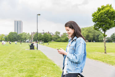 Lächelnde junge Frau in Jeansjacke, die im Park eine Textnachricht auf ihrem Smartphone schreibt - WPEF05699