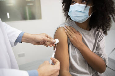 Doctor injecting COVID-19 vaccination to girl wearing protective face mask in medical room - PMF02212