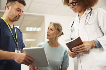 Ein Patient diskutiert mit einer Krankenschwester, die einen Tablet-PC mit einem Arzt in einem medizinischen Raum hält - PMF02219