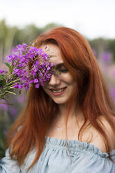 Smiling redhead woman with pink flowers at field - SSGF00444