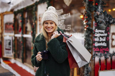 Lächelnde Frau mit Einkaufstüten und Weinbecher auf dem Weihnachtsmarkt - DAWF02414