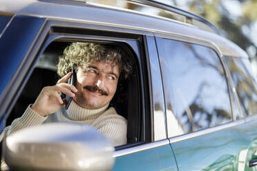 Lächelnder Mann, der mit einem Mobiltelefon spricht und aus dem Autofenster schaut - IFRF01366