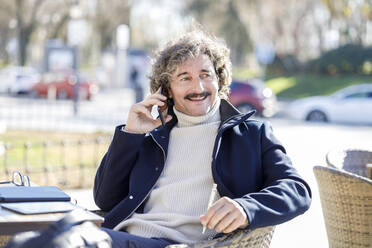 Lächelnder Mann, der in einem Straßencafé mit seinem Smartphone telefoniert - IFRF01374