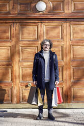 Man with shopping bags standing in front of wooden door on sunny day - IFRF01375