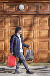 Smiling man walking with shopping bags in front of door on sunny day - IFRF01377