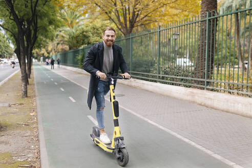 Fröhlicher Mann fährt E-Push-Roller auf dem Fahrradweg - JRVF02431