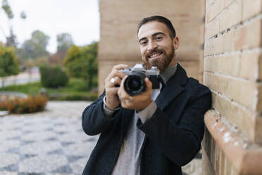 Happy young bearded man with camera leaning on wall - JRVF02440