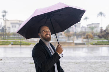 Fröhlicher junger Mann mit Regenschirm genießt den Regen - JRVF02441