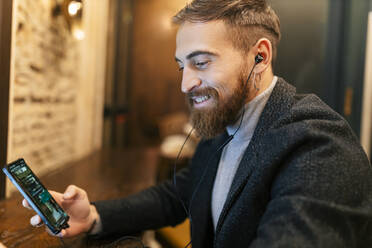 Happy young man using mobile phone and listening music through in-ear headphones - JRVF02450