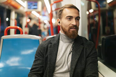 Young man looking out of tram window and listening music through in-ear headphones - JRVF02455