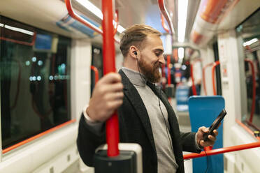 Man standing in tram using smart phone and listening music through in-ear headphones - JRVF02457