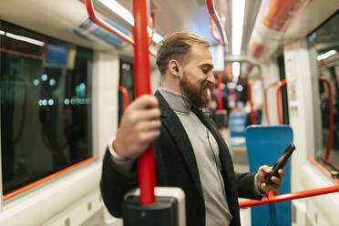 Mann steht in der Straßenbahn, benutzt sein Smartphone und hört Musik über In-Ear-Kopfhörer - JRVF02457