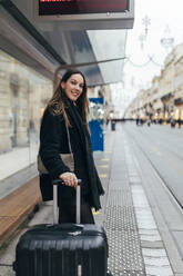 Smiling woman with suitcase waiting at tram station in city - JRVF02462