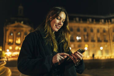 Lächelnde junge Frau, die in einer nächtlichen Stadt ein Smartphone benutzt - JRVF02483