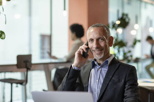 Lächelnder Geschäftsmann, der mit seinem Smartphone im Büro spricht - EIF03059