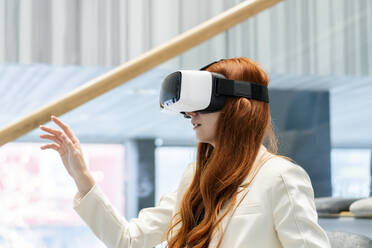 Redhead working woman wearing virtual reality headset gesturing in office - SSGF00451