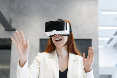 Aufgeregt arbeitende Frau, die einen Virtual-Reality-Simulator trägt und mit offenem Mund im Büro gestikuliert - SSGF00481