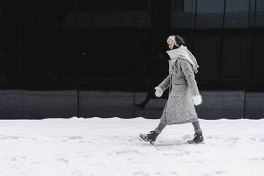 Frau in grauem Mantel geht auf verschneitem Fußweg an schwarzer Wand - SEAF00349