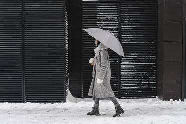 Nachdenkliche Frau mit Regenschirm auf dem Fußweg an der Mauer im Winter - SEAF00360