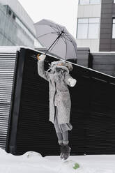 Fröhliche Frau mit Regenschirm springt auf verschneitem Fußweg - SEAF00376