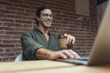 Lächelnder Geschäftsmann mit Einweg-Kaffeebecher und Laptop im Büro - JSRF01794