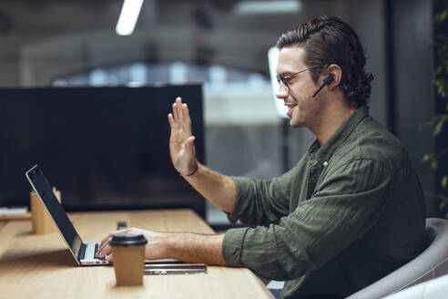Geschäftsmann mit winkender Hand bei einem Videoanruf in einem Coworking-Büro - JSRF01803