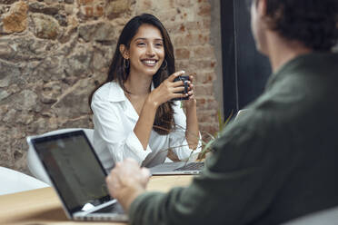 Smiling businesswoman holding coffee mug discussing with businessman at desk in office - JSRF01808