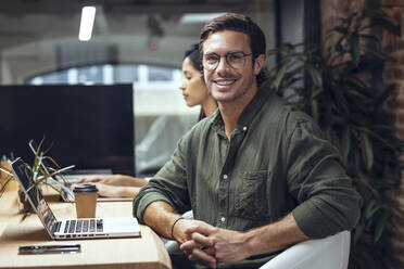 Selbstbewusster Geschäftsmann mit Laptop sitzt neben einem Kollegen am Schreibtisch im Büro - JSRF01810