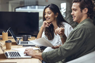 Smiling businessman and businesswoman discussing over documents and laptop at desk in coworking office - JSRF01813