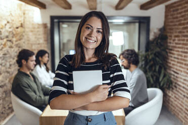 Lächelnde Geschäftsfrau mit Tablet-PC und Kollegen, die im Hintergrund im Büro arbeiten - JSRF01828