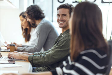 Lächelnder Geschäftsmann sitzt mit Kollegen am Schreibtisch in einem Coworking-Büro - JSRF01834