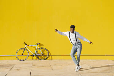 Glücklicher junger Mann tanzt vor einer gelben Wand an einem sonnigen Tag - JCZF00966