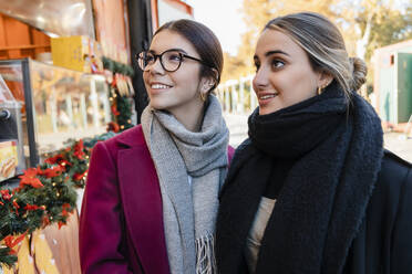 Smiling friends in warm clothing looking at store window - JRVF02498