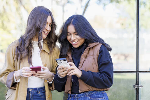 Lächelnde junge Frau teilt ihr Smartphone mit einem Freund auf dem Campus - IFRF01423