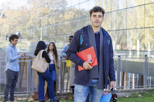 Junger Mann hält Akte mit Studenten im Hintergrund auf dem Campus - IFRF01443
