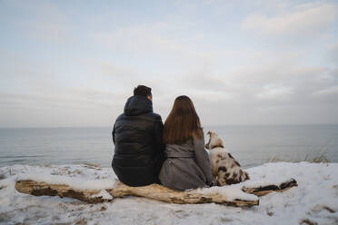 Young couple sitting with Australian Shepherd dog on snow covered log at beach - SSGF00494