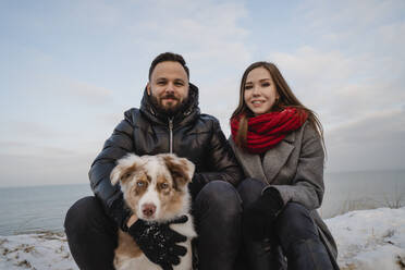 Lächelndes junges Paar sitzt mit Hund am Strand - SSGF00495