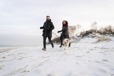 Junges Paar läuft mit Hund im Schnee bei Sonnenuntergang - SSGF00497