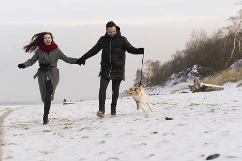 Glückliches junges Paar läuft mit Hund im Schnee bei Sonnenuntergang - SSGF00498